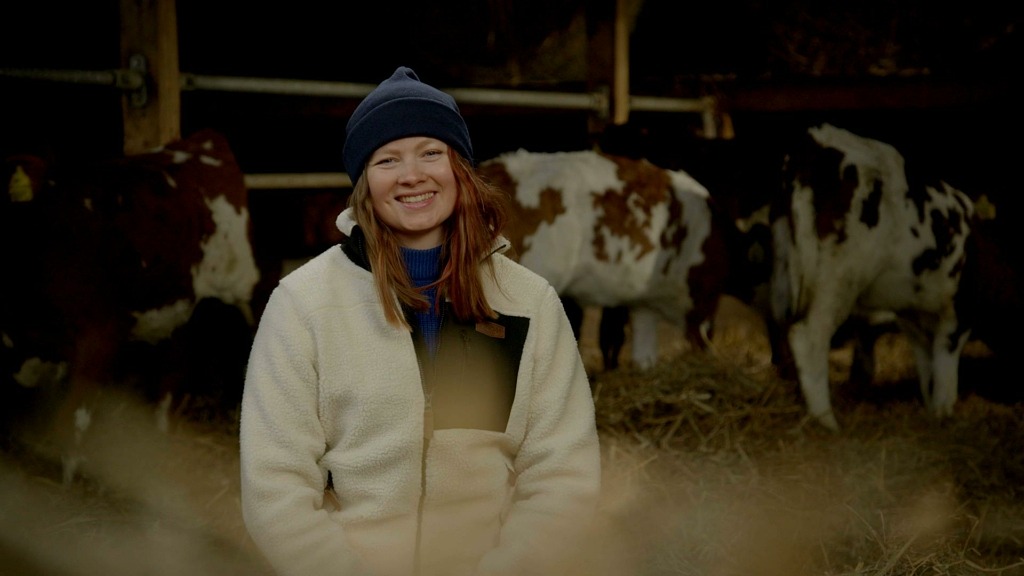 Alma Håkansson är en av mjölkbönderna i Bonde söker fru. Hon sitter med en vit jacka och blå mössa med kalvar i bakgrunden.