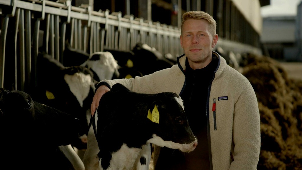 Isak Johansson är en av mjölkbönderna i Bonde söker fru. Han har en vit jacka och håller armen om en kalv.